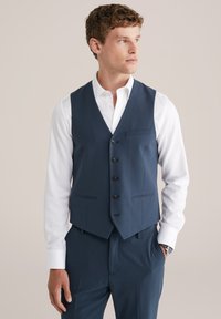 Mann in einer marineblauen Anzugweste und passender Hose mit einem weißen Hemd, blickt nach rechts mit einer Hand in der Tasche.