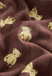 Brun tyg med en stickad textur och upprepade björnmotiv i ljust gult. Björnarna har runda ansikten och sitter upprätt.