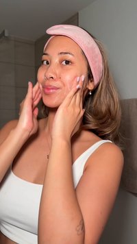 Ansiktsrens påført av en person som har på seg en lyserosa hårbånd og en hvit topp. Bakgrunnen har beige fliser og et håndkle.