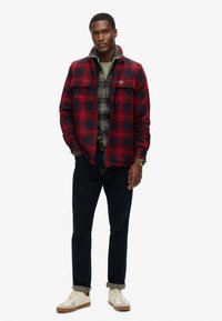 Rød og sort ternet flanneljakke med to brystlommer, lagret over en grønternet skjorte. Mørke jeans og cremefarvede sneakers fuldender outfittet.