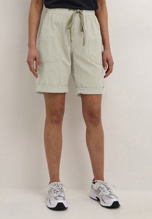 Person iført lyse beige knælange shorts med snøre og opsmøgede manchetter, kombineret med hvide atletiksko og en mørk top.