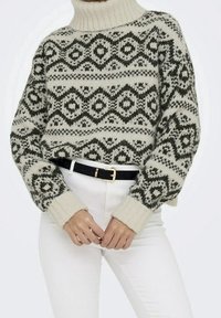 Pull à col roulé en maille crème avec motifs géométriques verts foncés ; poignets et ourlet côtelés ; coupe ample ; associé à une ceinture noire et un pantalon blanc.