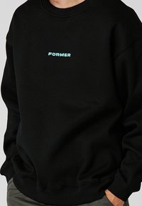 Sudadera negra con cuello redondo y puños de canalé. Presenta un pequeño logo bordado "FORMER" en color azul claro en la parte frontal. Material suave.