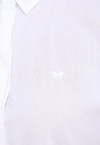 Camisa de algodón blanca con cuello abotonado y un pequeño logotipo bordado en blanco. La tela tiene una textura suave y ligera.
