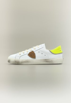Biały sneaker z wytartą podeszwą, brązową naszywką z boku, żółtym paskiem na pięcie, szarym wykończeniem i kremowymi sznurówkami, pokazany z profilu na neutralnym tle.