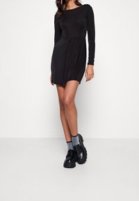 Robe noire à manches longues avec un haut ajusté et une jupe évasée. Le modèle porte des chaussures à plateforme noires et des chaussettes grises. Design simple et épuré.