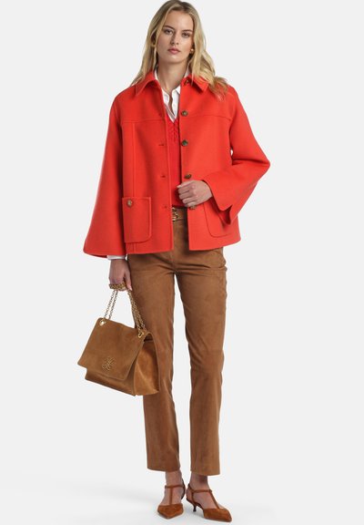 Ryškiai oranžinis vilnonis paltas su trimis sagomis, dviem priekinėmis kišenėmis ir plačiomis rankovėmis, derinamas su rudos spalvos zomšos kelnėmis ir atitinkančiais aukštakulniais.