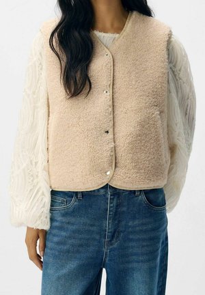 Woman wearing cream-colored sherpa snap-button vest over white textured long-sleeve blouse and blue denim jeans, cropped at shoulders and hips.
