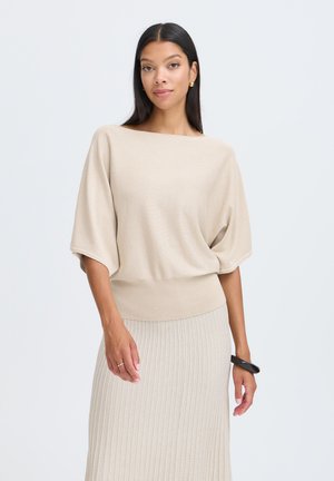Femme aux longs cheveux foncés portant un haut beige ample à manches courtes et une jupe en maille côtelée, accessoirisée avec des boucles d'oreilles dorées, une bague et un bracelet noir.