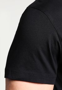 Close-up af en persons overarm iført en ensfarvet sort t-shirt med korte ærmer mod en neutral baggrund.
