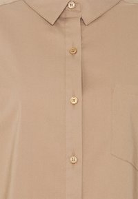 Chemise beige à boutons avec col pointu, trois boutons visibles, et une poche poitrine cousue sur le côté droit.
