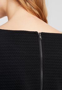 Mustekattu musta kangas, jossa on siksak-kuvio, ja takana näkyvä metallinen vetoketju. Pehmeä kaulus, jossa on hieman kaarevuutta.