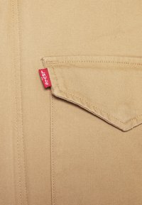 Beige skjorta i tyg med en spetsig ficka som har en röd Levi's-flik på sömmen. Texturerad yta med synliga sömnadsdetaljer.