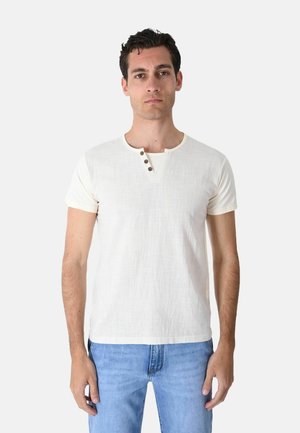 Uomo che indossa una camicia crema a maniche corte con tre bottoni vicino al colletto e jeans azzurri, in piedi davanti a uno sfondo semplice.