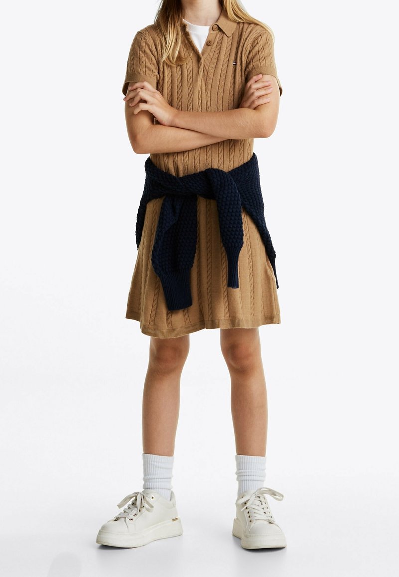 Personne debout, les bras croisés, portant une robe en maille torsadée beige, des baskets blanches et un pull bleu marine noué autour de la taille.