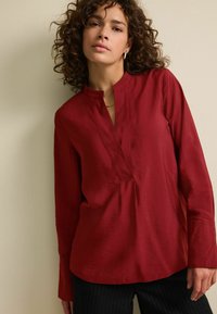 Červená košile s dlouhým rukávem a mandarinovým límcem, s částečnou légou. Hladký materiál s mírným leskem. Volný střih s zakřiveným lemem.