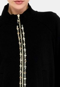 Giacca nera con zip e colletto alto, dotata di perline decorative lungo la zip e tasche laterali. Tessuto morbido.