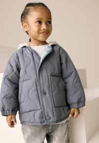 Veste grise matelassée avec capuche, dotée de deux poches avant, d'une doublure intérieure bleue et de motifs de coutures arrondis. Portée avec un jean gris.