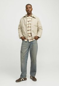 Beige bomberjakke over en ternet skjorte i creme og brun, parret med lyseblå straight-leg jeans og brune sneakers med hvide striber.