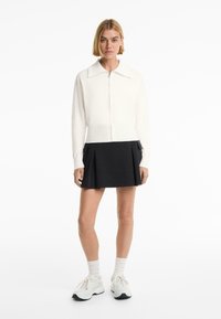 White zip-up sweater with a collar above a black pleated skirt. The outfit is paired with light sneakers and white ribbed socks.