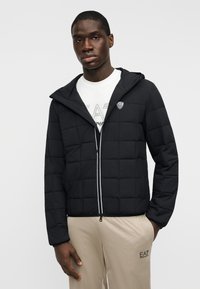 Μαύρο μπουφάν με πούπουλο, με φερμουάρ εμπρός, κουκούλα και λευκές κορδόνια. Διαθέτει λογότυπο στο αριστερό στήθος και μακριά μανίκια.