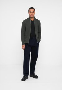 Pull en laine vert foncé zippé, T-shirt noir, pantalon bleu marine et chaussures noires. Texture lisse, coupe ajustée et détails minimalistes.