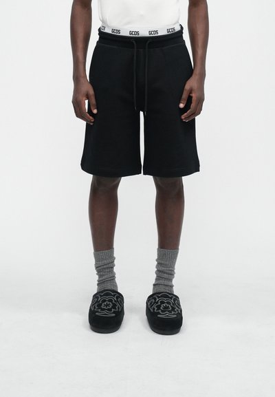 Schwarze Baumwollshorts mit elastischem Bund, verziert mit einem weißen Logo, kombiniert mit grauen gerippten Socken und schwarzen Slippern mit floraler Stickerei.