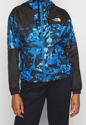 Osoba ubrana w niebiesko-czarną, wzorzystą kurtkę z kapturem North Face z czarnymi rękawami i czarne spodnie na jednolitym tle.