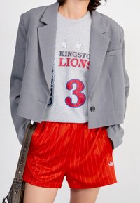 Grå skräddarsydd kavaj, grå t-shirt med tryck "KINGSTON LIONS 3" och röda kortbyxor med smala ränder och Adidas-logga.