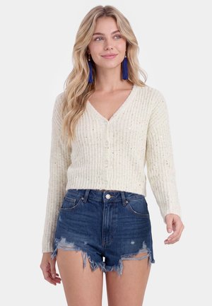 Junge Frau mit langen blonden Haaren, die einen weißen, geknöpften Cardigan, blaue Quasten-Ohrringe und zerstörte hoch taillierte Jeansshorts trägt.
