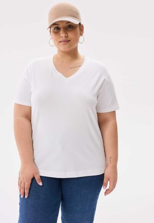 T-shirt blanc en V, fabriqué en tissu doux, avec des manches courtes. Associé à un jeans bleu. Le modèle porte une casquette beige et des boucles d'oreilles créoles.