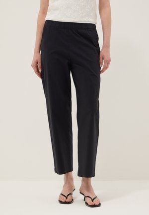 Mujer con pantalones negros de pierna recta con cinturilla elástica, camiseta sin mangas blanca con textura y sandalias negras con tacón.