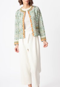 Jakna s uzorkom od zelene i krem tkanine, s narančastim obrubom i dekorativnim gumbima. Uparena s laganim, širokim hlačama svijetle boje.