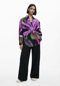 Camicia a maniche lunghe con un vivace motivo floreale viola e verde su sfondo nero; abbinata a pantaloni neri a gamba larga.