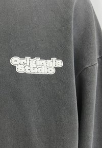 Šedá mikina s velkým, vyvýšeným logem "Originals Studio" v bílé barvě. Materiál má texturovaný, měkký povrch.