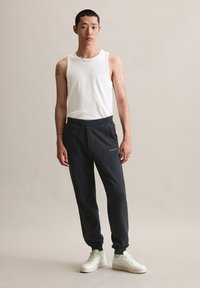 Weißes ärmelloses Tanktop, graue Jogginghose mit Fronttasche, dezente Markenkennzeichnung und weiße Turnschuhe. Glatter Stoff, lässige Passform und modernes Design.
