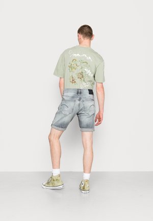 Homme portant un t-shirt graphique vert clair, un short en denim gris retroussé et des baskets montantes vertes, debout devant un fond blanc.
