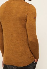 Pull en maille couleur jaune moutarde avec manches longues et coupe ajustée. Présente un petit patch logo sur le haut du bras. Texture douce.