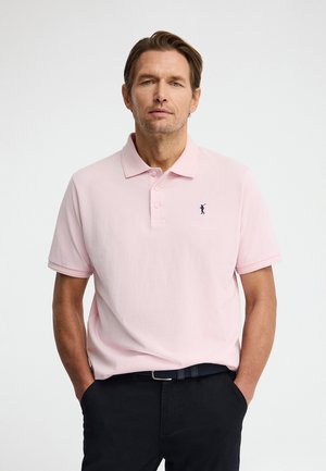 Hombre con cabello castaño claro que lleva una camisa tipo polo rosa claro con un pequeño logo de golfista en azul oscuro, pantalones negros y un cinturón oscuro, con las manos en los bolsillos.