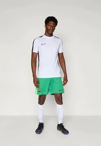 Άνδρας αθλητής φορώντας λευκό και μαύρο αθλητικό μπλουζάκι, πράσινες σορτς με λογότυπο της ομάδας, λευκές κάλτσες μέχρι το γόνατο και μαύρα ποδοσφαιρικά παπούτσια, στέκεται μπροστά σε απλό φόντο.