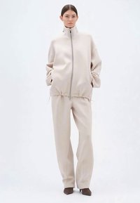 Lys beige oversized jakke med høy krage, glidelås og to lommer, sammen med matchende vide bukser og brune hæler.