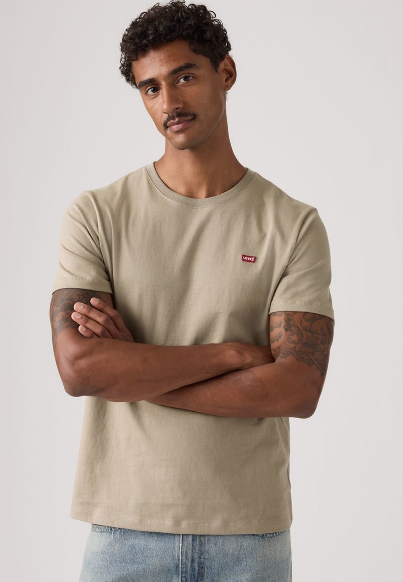 Beige bomullst-shirt med korta ärmar, rund halsringning och en liten röd Levi's-logga på vänster bröst, bärs med ljusblå denimjeans.