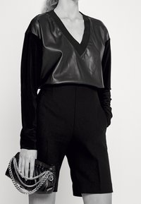 Schwarzes Langarmoberteil mit V-Ausschnitt aus glattem Material, kombiniert mit maßgeschneiderten schwarzen Shorts. Eine Hand hält eine schwarze Clutch mit Kettenakzenten.