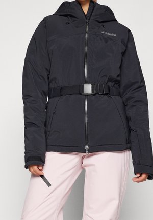 Persoon draagt een zwarte winterjas met ritssluiting en riem en lichtroze broek, houdt een zwart rechthoekig voorwerp vast in de linkerhand.