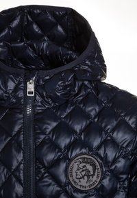 Granatowa pikowana kurtka puchowa z zamkiem błyskawicznym, z okrągłym, haftowanym logo na piersi i kapturem. Gładka, błyszcząca tekstura.