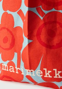 Czerwona i niebieska bawełniana torba z dużymi kwiatowymi wzorami o teksturowanym wykończeniu oraz nazwą marki "marimekko" w białym napisie.
