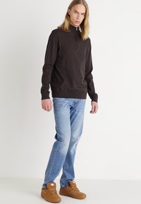 Suéter de manga larga marrón oscuro con cierre de cuarto, combinado con jeans azul claro y zapatillas marrones, de pie contra un fondo neutro.