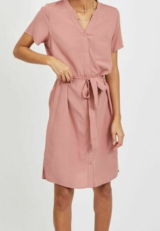 Femme ajustant la ceinture d'une robe rose à manches courtes, longueur genou, boutonnée, avec une poche poitrine, sur un fond uni.