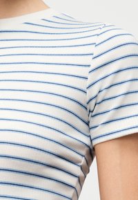 Chemise blanche à manches courtes côtelée avec de fines rayures horizontales bleues portée sur le torse et le bras.