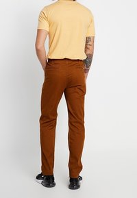 Brązowe bawełniane spodnie o slim fit z dwiema tylnymi kieszeniami, w połączeniu z żółtą koszulą z krótkim rękawem, widoczne od tyłu.